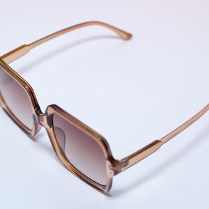 May include: Square-framed sunglasses with a light brown frame and gradient brown lenses. The sunglasses have a classic design with a modern twist, suitable for various face shapes. The temples are a lighter shade of brown.