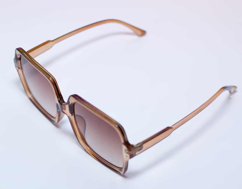 May include: Square-framed sunglasses with a light brown frame and gradient brown lenses. The sunglasses have a classic design with a modern twist, suitable for various face shapes. The temples are a lighter shade of brown.