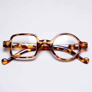 May include: Eyeglasses with a unique design. One lens is square, the other is round. The frames are a tortoiseshell pattern with a brown and yellow color scheme. The arms of the glasses have a heart-shaped design at the end.