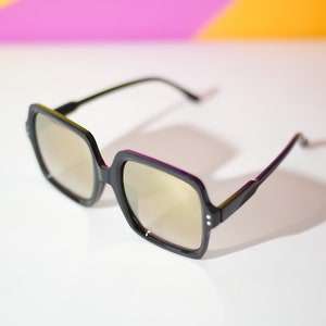 May include: Black square-framed sunglasses with gold-tinted lenses. The sunglasses have a thick frame and a modern design. The background is white with a gradient of pink, purple, and orange.