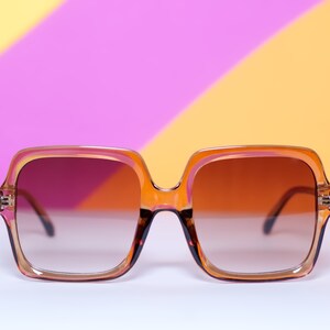 May include: Oversized square sunglasses with a gradient brown tint. The frames are a translucent orange-brown color with a square shape. The sunglasses are set against a colorful background of pink, yellow, and orange.