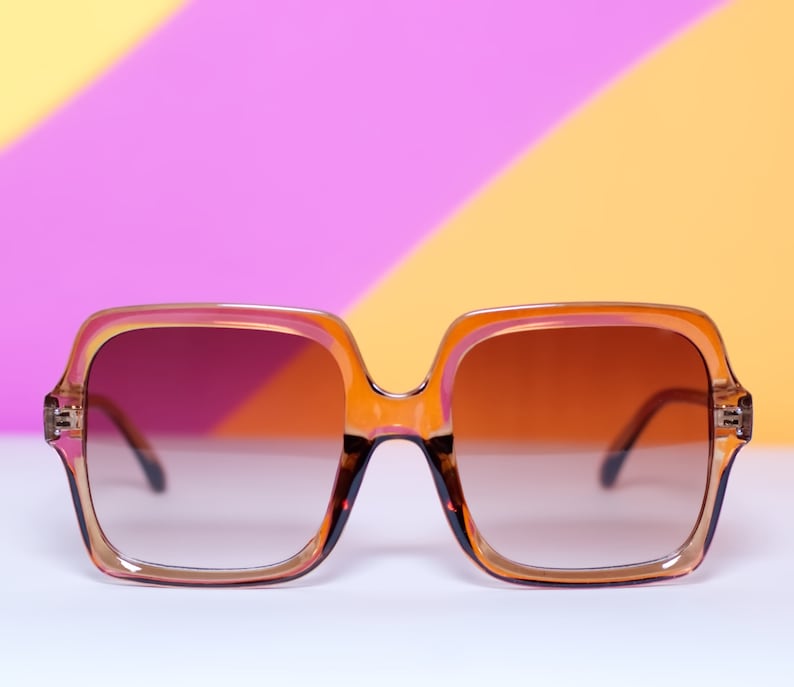 May include: Oversized square sunglasses with a gradient brown tint. The frames are a translucent orange-brown color with a square shape. The sunglasses are set against a colorful background of pink, yellow, and orange.