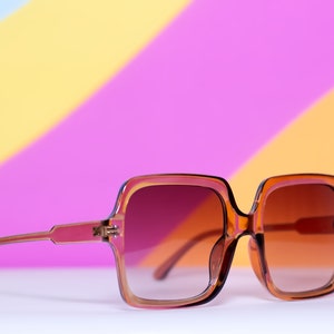 May include: Brown square-framed sunglasses with gradient lenses. The frames are a translucent amber color, and the lenses transition from a dark brown at the top to a lighter shade at the bottom. The sunglasses are set against a colorful background.