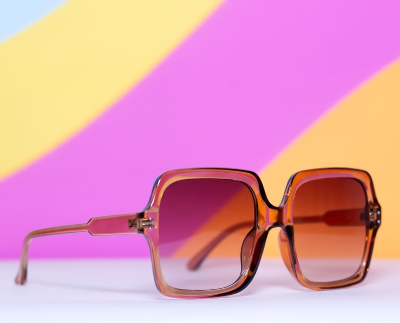 May include: Brown square-framed sunglasses with gradient lenses. The frames are a translucent amber color, and the lenses transition from a dark brown at the top to a lighter shade at the bottom. The sunglasses are set against a colorful background.