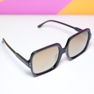 May include: Black square-framed sunglasses with brown tinted lenses. The sunglasses have a thick frame with small silver accents on the front. The arms are thin and black. The background is white with a gradient of pink, yellow, and purple.
