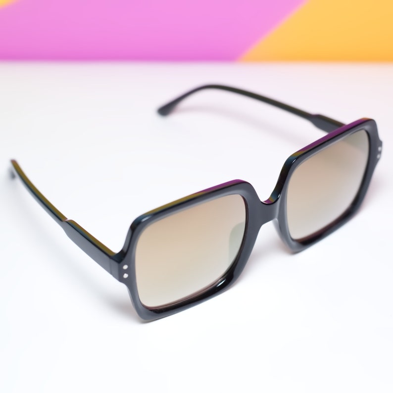 May include: Black square-framed sunglasses with brown tinted lenses. The sunglasses have a thick frame with small silver accents on the front. The arms are thin and black. The background is white with a gradient of pink, yellow, and purple.