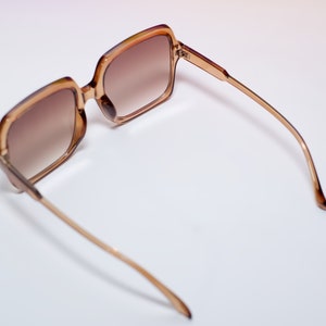 May include: Brown-tinted sunglasses with a square frame and thin, gold-colored arms. The lenses have a gradient tint, transitioning from a darker shade at the top to a lighter shade at the bottom. The sunglasses are set against a white background.