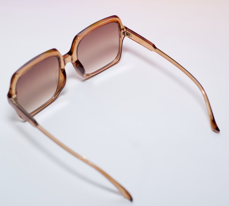 May include: Brown-tinted sunglasses with a square frame and thin, gold-colored arms. The lenses have a gradient tint, transitioning from a darker shade at the top to a lighter shade at the bottom. The sunglasses are set against a white background.