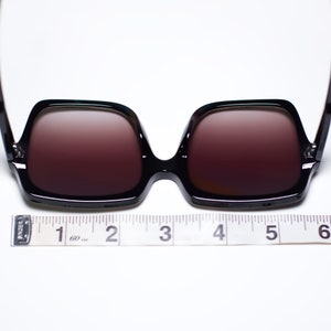 May include: Black square-framed sunglasses with brown-tinted lenses. The sunglasses are measured with a tape measure, approximately 6 inches wide. The frames are glossy and the lenses are a gradient color.