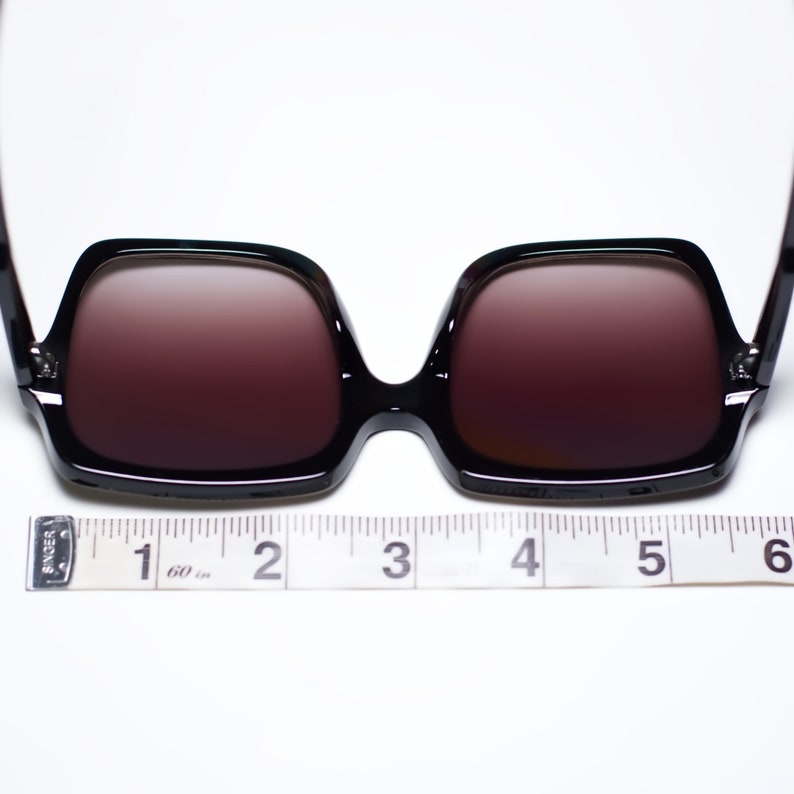 May include: Black square-framed sunglasses with brown-tinted lenses. The sunglasses are measured with a tape measure, approximately 6 inches wide. The frames are glossy and the lenses are a gradient color.