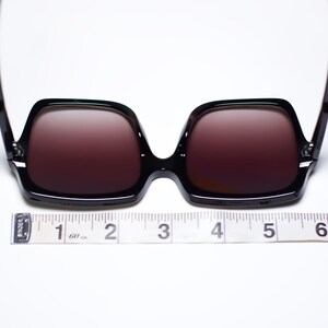 May include: Black square-framed sunglasses with dark lenses. The sunglasses are measured with a ruler, approximately 5 inches wide. The frames are glossy and the lenses have a gradient tint.