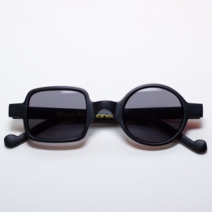 May include: Black sunglasses with a unique design. One lens is square, the other is round. The frames are matte black with the text "Bold For" and "onec" in gold. The arms have a curved design.
