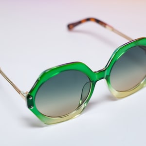 May include: Green and gold-toned sunglasses with a unique hexagonal frame. The lenses transition from a dark to a lighter shade. The temples are gold with tortoise shell tips. A stylish accessory for eye protection.