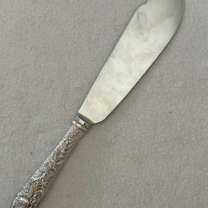 May include: A silver-toned serving knife featuring a wide, smooth, curved blade and a decorative handle embossed with intricate floral and leaf patterns. The metal handle displays a detailed relief design of various blossoms and foliage throughout.