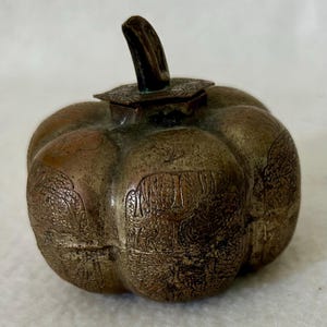 Puede incluir: Un recipiente en forma de calabaza de color bronce con tapa y tallo. El recipiente tiene patrones florales detallados grabados en su superficie. La tapa es cuadrada con un pequeño tallo. El diseño general es antiguo y decorativo.