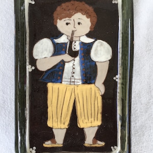 Op de afbeelding: Een keramische tegel met een groene rand en een bruine achtergrond. Een cartoonillustratie van een persoon met bruin haar die een blauwe vest, een wit shirt en een gele broek draagt, houdt een pijp in zijn mond.
