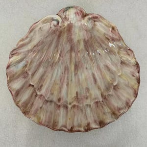May include: A ceramic seashell dish with a pink, white, and brown glaze. The dish is shaped like a scallop shell and has a slightly raised rim.