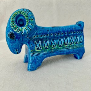 May include: A ceramic ram figurine in shades of blue and green. The ram has large curved horns, a detailed face, and a decorative pattern along its back. The figurine is standing on four legs and has a small tail. The overall design is artistic and decorative.