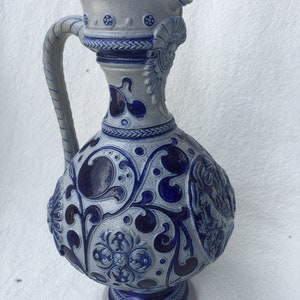 Op de afbeelding: Een blauwe en witte keramische kan met een bloemenmotief en een handvat. De kan heeft een rond lichaam en een hoge, slanke hals.