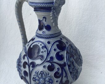 Puede incluir: Una jarra de cerámica azul y blanca con un diseño floral y un asa. La jarra tiene un cuerpo redondo y un cuello alto y delgado.