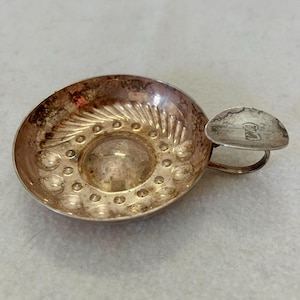 Puede incluir: Un pequeño plato circular de metal plateado con una sola asa. El interior presenta una cúpula central elevada rodeada por un anillo de hendiduras circulares en relieve y un patrón en forma de abanico en el borde superior. Un sello es visible en el asa.
