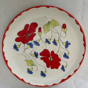 Puede incluir: Un plato de cerámica blanco con un borde rojo festoneado. El plato está decorado con flores rojas, flores azules y hojas verdes. Las flores están pintadas con un diseño estilizado.