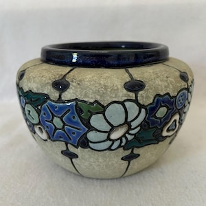 May include: A round ceramic vase with a speckled beige exterior and a dark blue rim. The vase is decorated with a floral pattern in shades of blue, green, and white, with black accents.