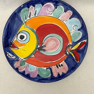 May include: A hand-painted ceramic plate with a vibrant fish design. The fish features yellow, orange, and red colors, with a blue background. The plate has a whimsical, artistic style, perfect for kitchen decor.
