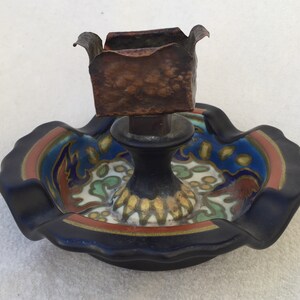 May include: An antique candle holder with a black base and a decorative, colorful dish. The dish features a blue, red, and green floral design. The candle holder itself is topped with a copper-colored square cup.