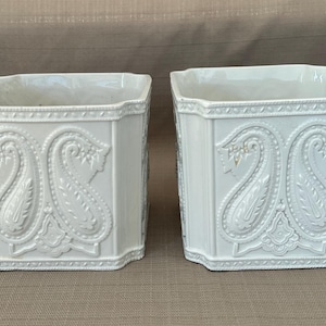 May include: Two white ceramic planters with a square shape and a detailed paisley design. The planters have a textured surface and are suitable for indoor or outdoor use.