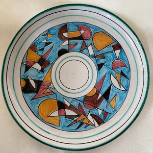Puede incluir: Un plato redondo de cerámica con fondo azul y un diseño geométrico en tonos marrones, amarillos, rojos y negros. El plato tiene un borde verde y rayas blancas.