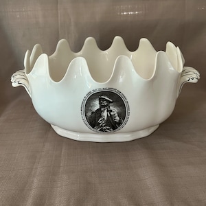 Puede incluir: Tazón de cerámica blanco con borde festoneado y dos asas. El tazón presenta una imagen en blanco y negro de un hombre con sombrero con el texto "I PREFER NO DESSERT THAT YOU MAY COMPARES SUCH EXCELLENT BREAD AND CHEESE WITH THE FINE FRUIT OF JAMAICA".