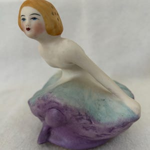 May include: A porcelain figurine of a woman with blonde hair, blue eyes, and rosy cheeks. The figurine is wearing a dress with a purple and teal color scheme. The woman is in a seated position, with her arms extended.