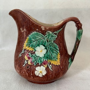 May include: A brown ceramic creamer with a leaf and flower design. The creamer has a handle that is shaped like a branch.