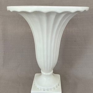 Pode incluir: Um vaso de cerâmica branco com um design canelado e uma base quadrada. O vaso tem uma abertura larga e um pescoço estreito.