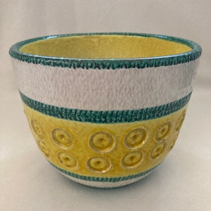 Puede incluir: Un macetero de cerámica blanco y amarillo con un borde verde y un patrón de círculos alrededor del centro.
