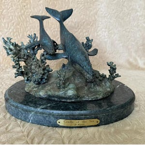 May include: A bronze sculpture of a mother and baby whale swimming through coral. The sculpture is on a green marble base with a gold plaque that reads "Cradle of the Sea" by J. Wright.