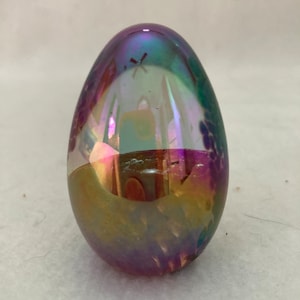 May include: A iridescent glass egg with a purple, blue, and green color scheme. The egg has a white interior with a faint image of a building.