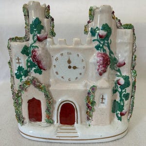 Puede incluir: Reloj de cerámica con forma de castillo, con una esfera en el centro. El castillo está pintado en un tono rosa claro con puertas rojas y vides decorativas con uvas rosas. Las manecillas del reloj señalan las 12 en punto.
