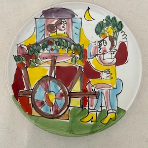 May include: A hand-painted ceramic plate featuring a whimsical scene of a person with a fruit cart. The plate has a white background with a green border. The cart is red, yellow, and blue, and the person is wearing yellow and red clothing.