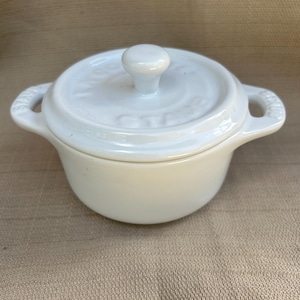 Könnte beinhalten: Weiße Keramik-Auflaufform mit Deckel und zwei Griffen. Die Form ist klein und rund, und der Deckel hat einen Knopf oben.
