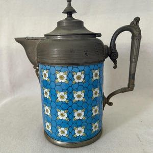 May include: A cylindrical metal pitcher featuring a bright blue enamel body with a repeating pattern of white flowers and a dark grey honeycomb grid. The pitcher includes a grey metal lid with a finial, a curved spout, and a decorative handle.