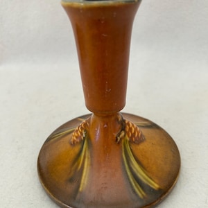 May include: A glazed ceramic candlestick holder featuring a warm brown finish. The base is decorated with a raised, painted motif of pinecones and green pine needles. The stem is cylindrical with a flared top opening for a candle.