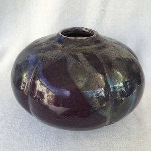May include: A round, dark purple ceramic vase with a glossy finish. The vase has a subtle iridescent sheen and a slightly textured surface.