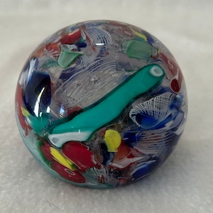 May include: A clear glass paperweight with a variety of colorful glass pieces embedded inside. The paperweight is round and has a smooth surface. The colors include red, blue, green, yellow, and white.