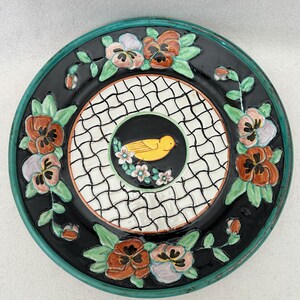 May include: Decorative ceramic plate with a black background and a teal rim. The plate features a central design of a yellow bird surrounded by white flowers. The outer edge is decorated with colorful pansies and green leaves.