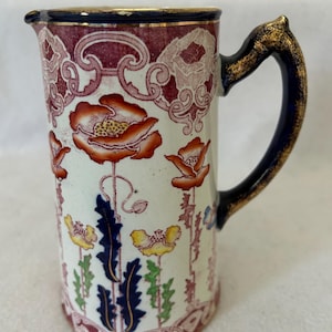 May include: A ceramic pitcher with a white base, adorned with red poppies and floral patterns. The rim and handle are accented with gold and dark blue. The pitcher's design includes a decorative border and a dark blue handle.