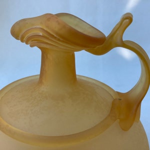 May include: A frosted glass pitcher with a unique, curved handle and a wide, round base. The pitcher is a pale yellow color.