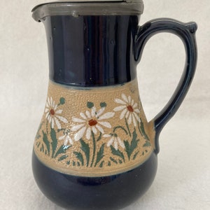 May include: A blue and brown ceramic pitcher with a pewter lid and a floral design. The pitcher has a white daisy pattern with green leaves.
