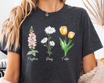 May include: A dark gray t-shirt with a graphic of three flowers: a foxglove, a daisy, and a tulip. The flowers are illustrated in a watercolor style with pink, white, and yellow colors. The names of the flowers are written below each flower.